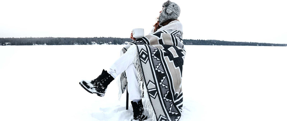 a man is is sitting in the snow wrapped in blanket with cup of coffee in the hand